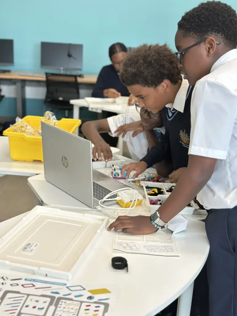 Three students in school uniforms work together at a table with a laptop and robotics kit, assembling pieces and consulting instructions in a classroom with computers in the background.