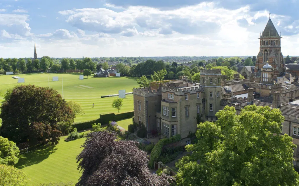 A large historic building with towers, part of the renowned Rugby School, sits beside a well-maintained sports pitch and lush green trees under a partly cloudy UK sky.