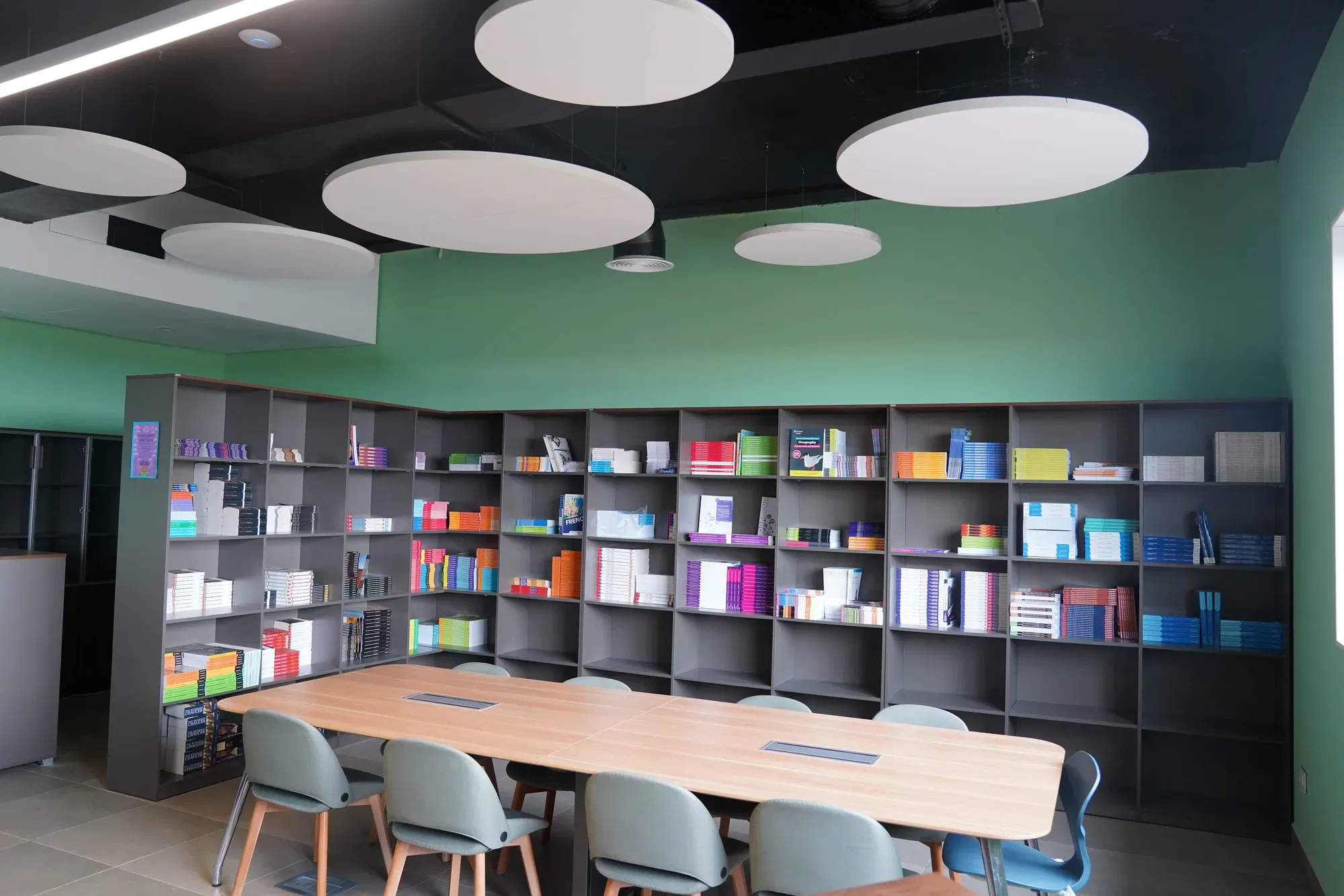 A modern meeting room with a long wooden table, eight teal chairs, green walls, a bookcase filled with colourful books and boxes, and large round white ceiling panels.