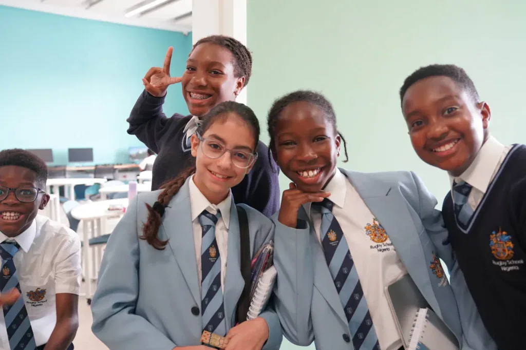 Five smiling students in school uniforms pose together in a classroom with teal walls. One student flashes a peace sign, whilst others hold exercise books and stand closely, appearing cheerful and friendly.