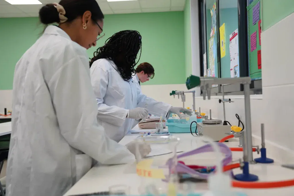 Three students in white lab coats and gloves work on experiments at a laboratory bench, surrounded by scientific equipment and colourful papers on the wall.