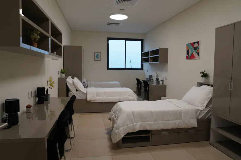Modern, tidy dormitory room with two single beds, desks, chairs, and shelves. Neutral colours, large window, minimalist decor, and plants create a clean, organised atmosphere.