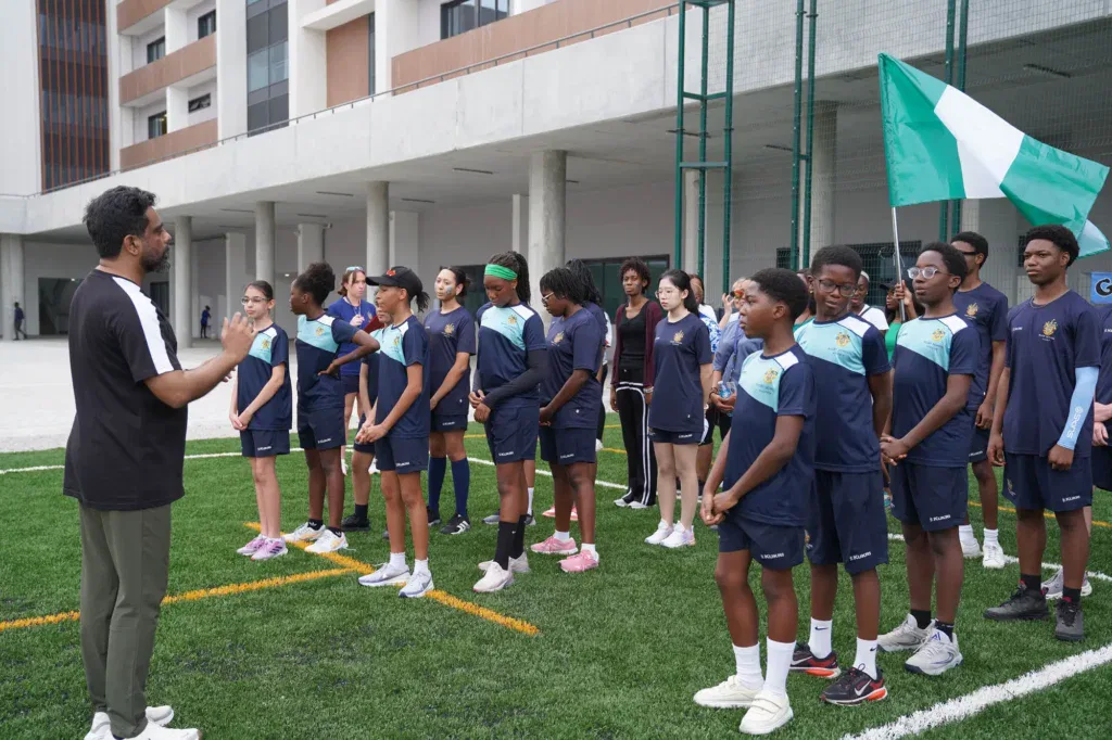 A group of students in sports kit stand in rows on a grass pitch, listening to an adult coach. A large green and white flag is held in the background near a modern building.
