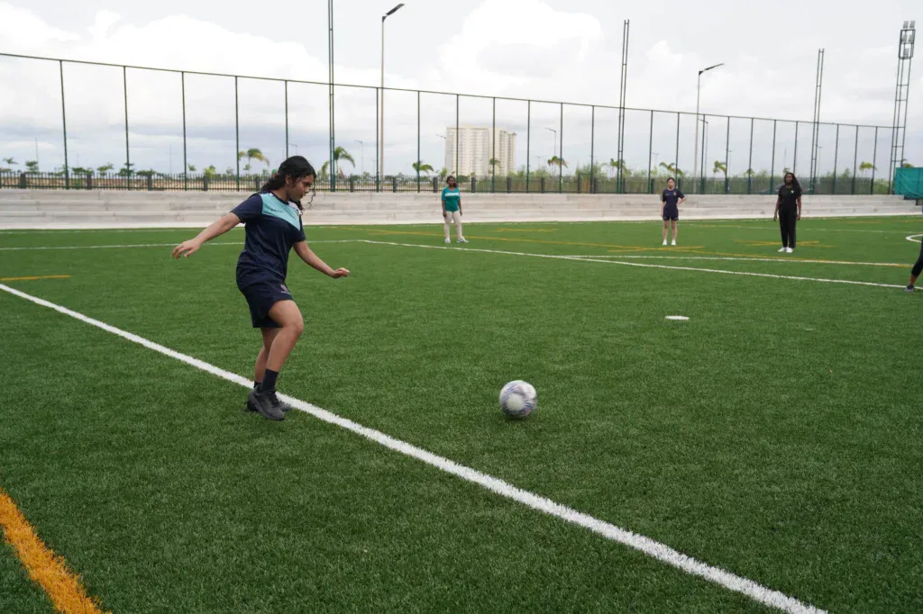 A young person in sports kit is about to kick a football on a green outdoor pitch, while four others stand in the background near the goal line.