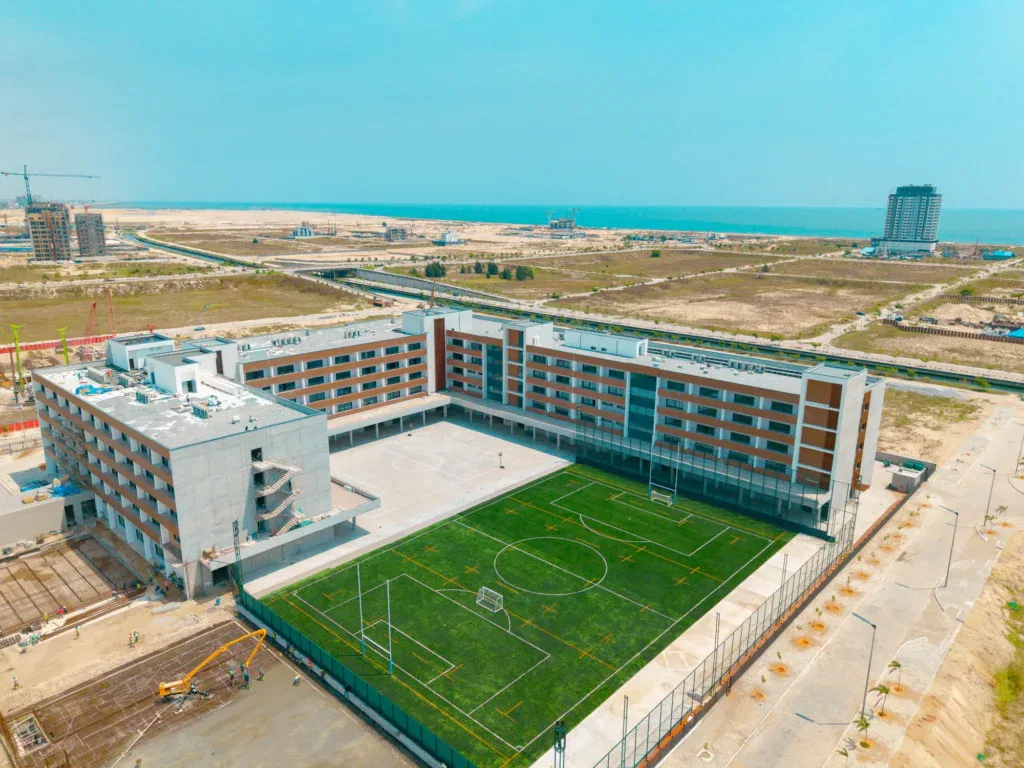 Aerial view of a large, modern school building with a green sports pitch in the centre, surrounded by paved areas and construction sites, near a coastline with distant buildings and open land.