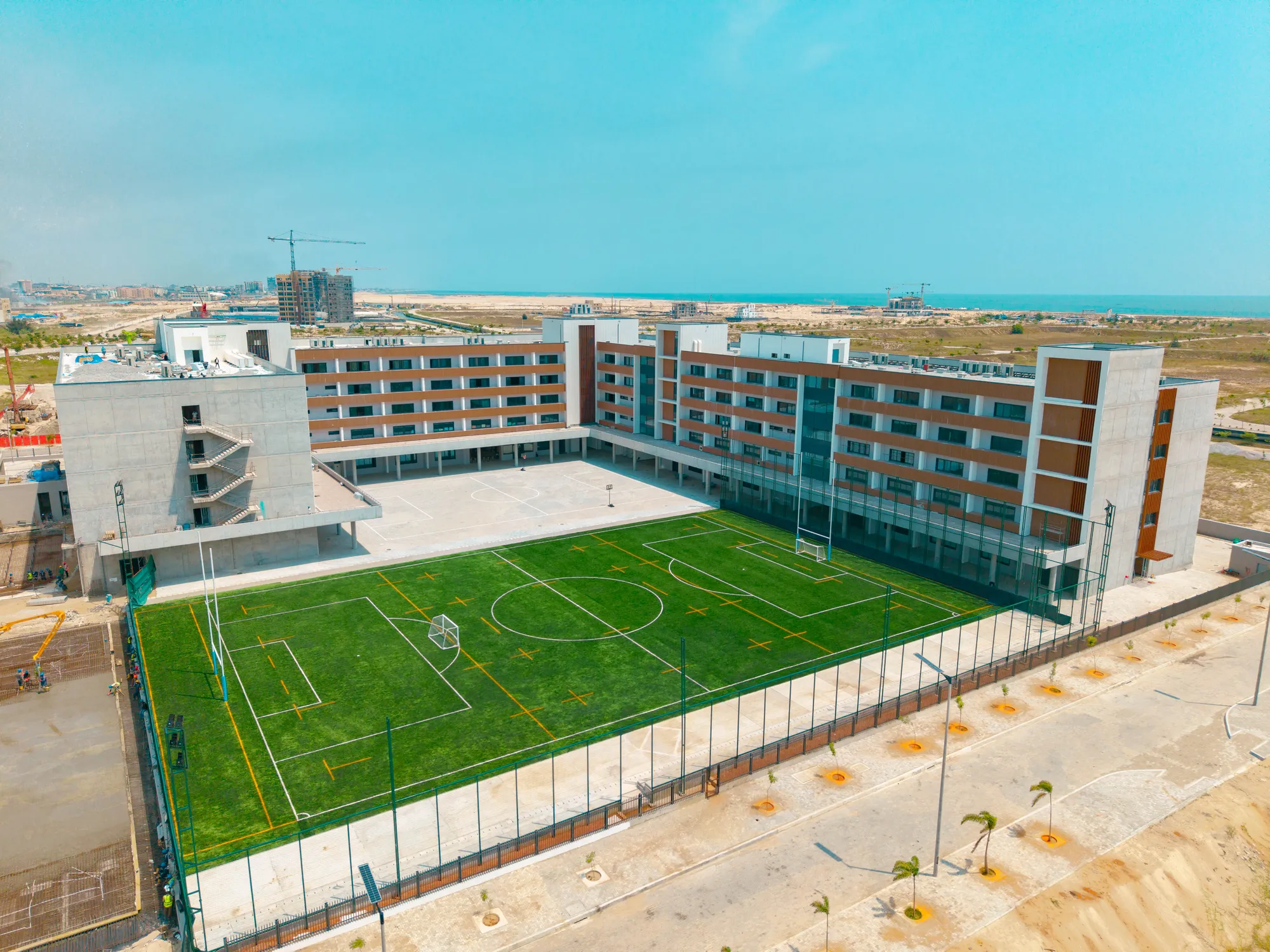A modern school building with four storeys surrounds a large, fenced, artificial turf football pitch. The area is clear and bright, with coastal scenery and the sea in the background.