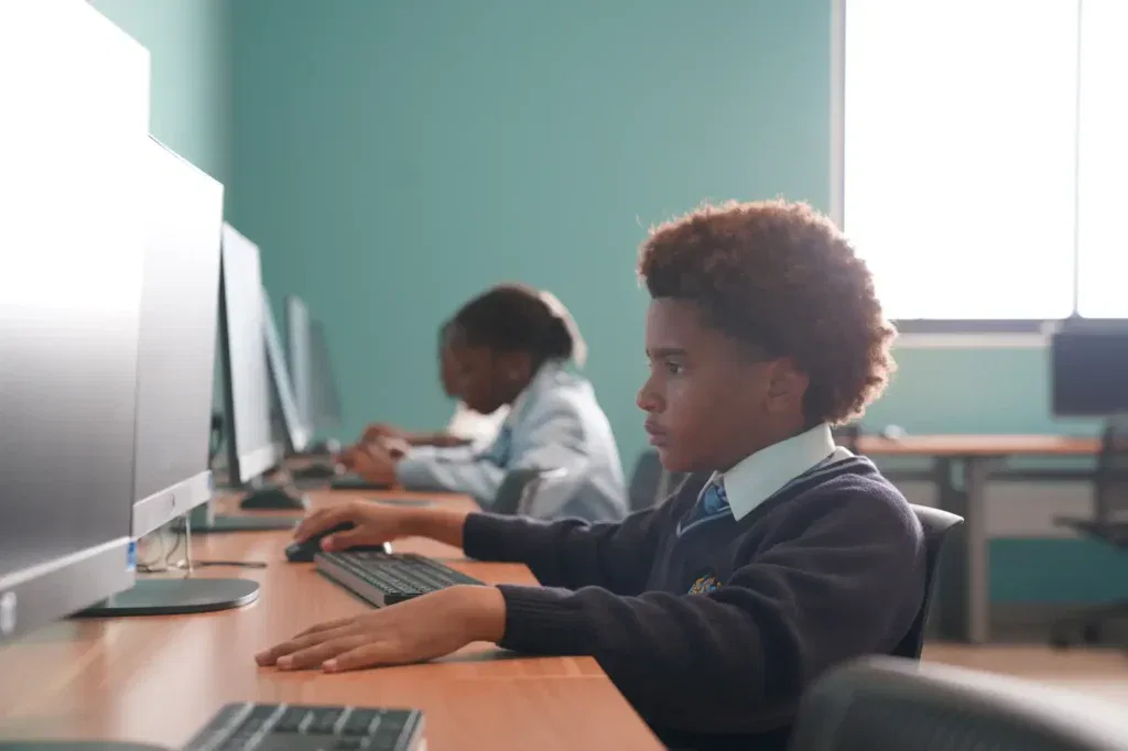Two students in uniforms work on desktop computers in a classroom with teal walls, engaging with the Lower School Curriculum. The pupil in the foreground is focused on the screen.