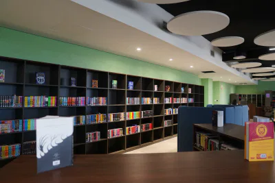 Modern library interior with long black bookshelves filled with colourful books against a green wall, overhead circular ceiling panels, and pamphlets displayed on dark wooden tables. Register now to explore this vibrant reading space.